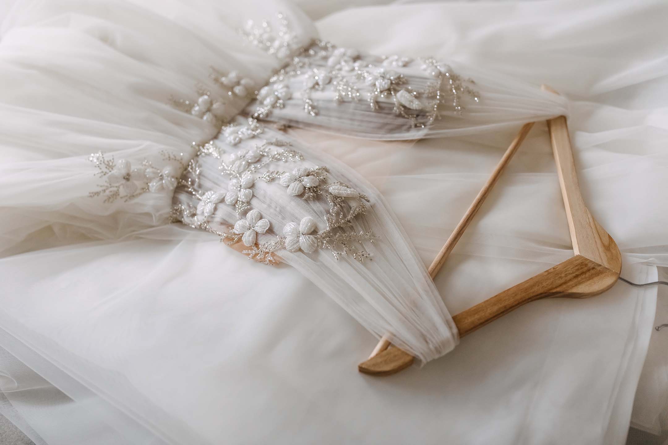 A white floral sheer wedding dress on it's hanger laying over fabric.